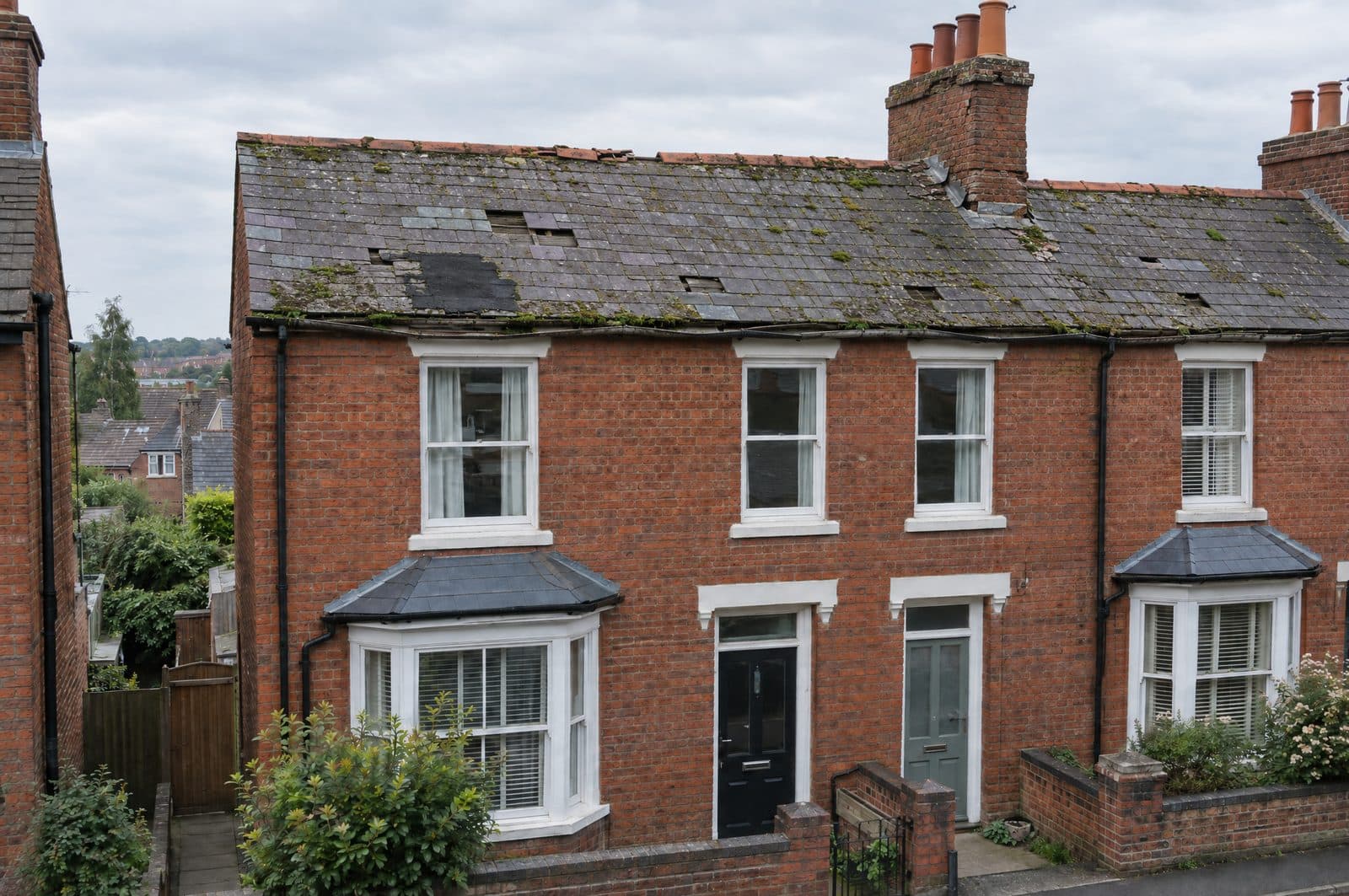 A terraced house with a worn slate roof: slipped and missing slates, moss, a black patch repair and a broken ridge