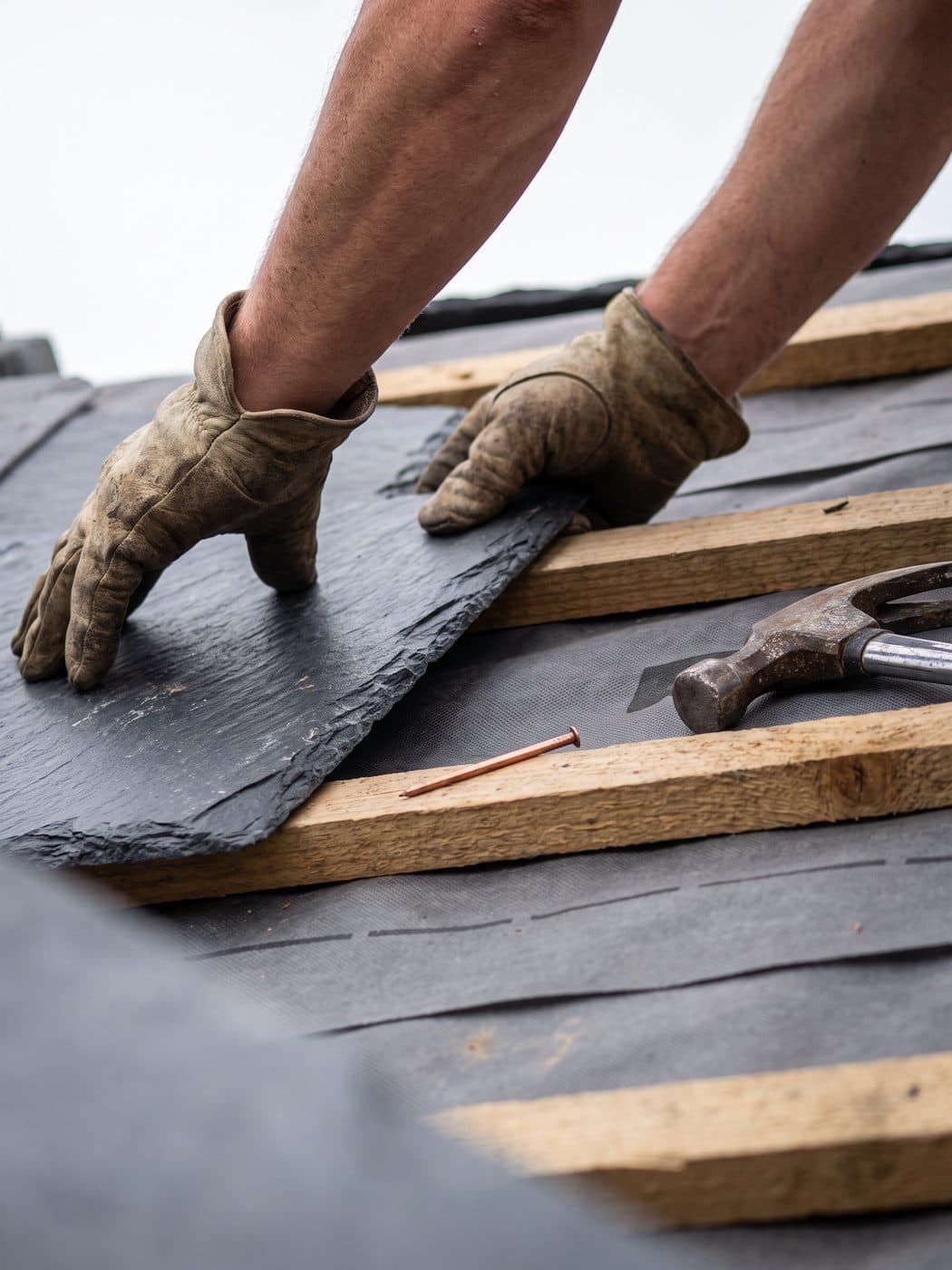 Close view of gloved hands positioning a natural slate onto timber battens, with copper nails and membrane visible beneath