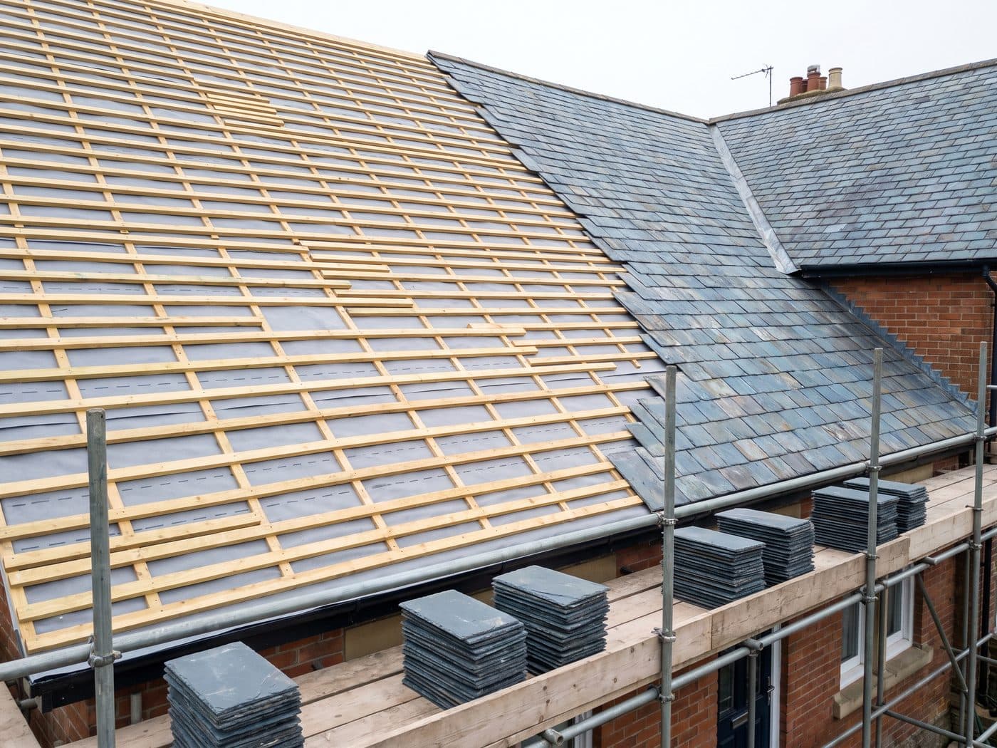A terraced house roof stripped back to new battens over breathable membrane, with old slate remaining on one side and new slates stacked on the scaffold