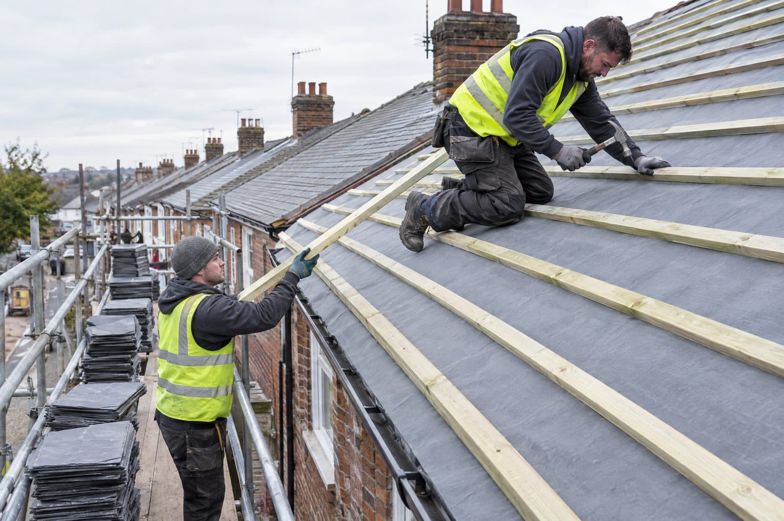 Two roofers fixing new timber battens over breathable membrane on a stripped terraced-house roof, with new slates stacked on the scaffold