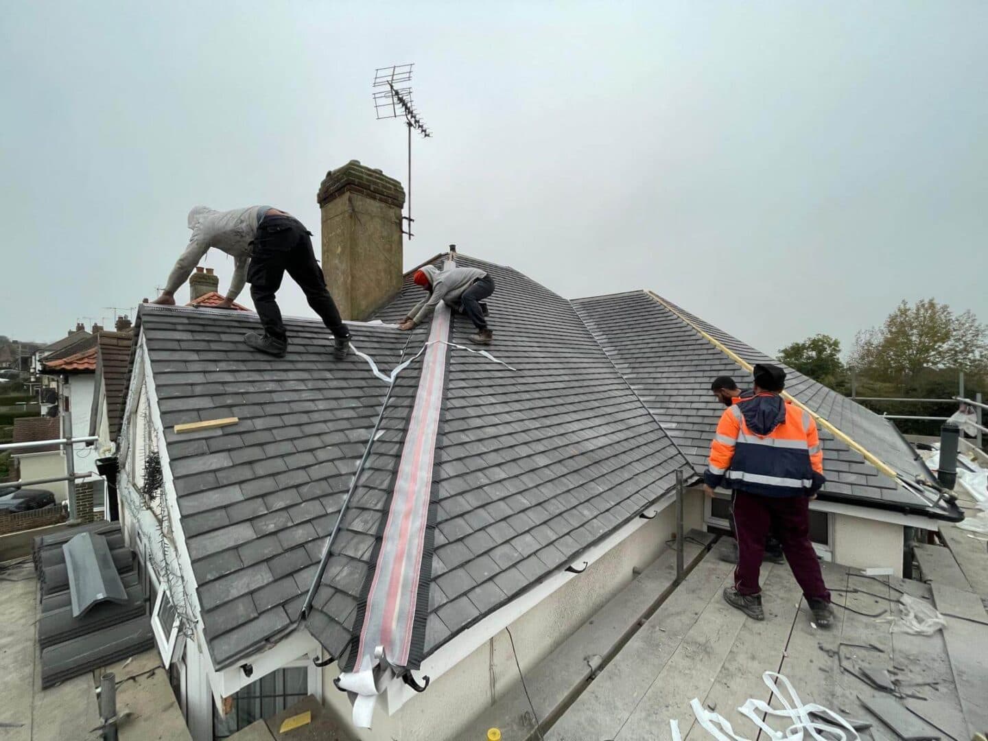 Three roofers on a grey tiled hip roof, one unrolling a dry hip roll along the hip line