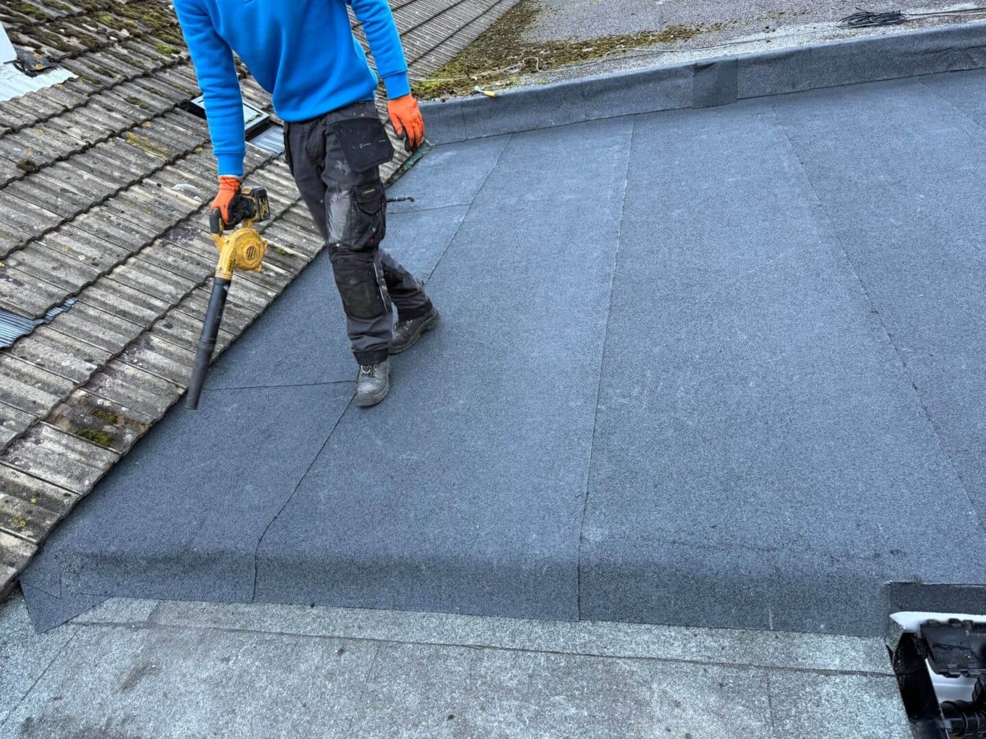 A roofer clearing a new mineral-felt flat roof with a blower, beside an older tiled roof